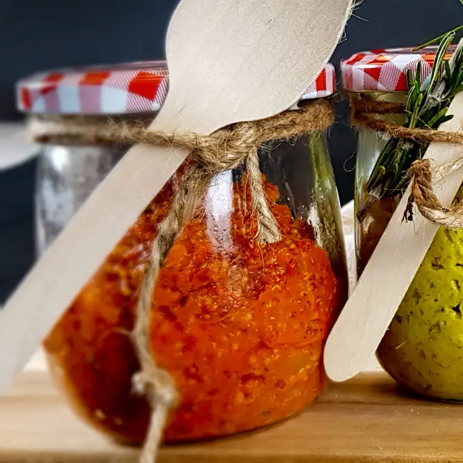 Kräftig roter Aufstrich aus Paprika und Quinoa, in einem schönen Einmachglas mit Kräutern und einem Holzlöffel dekoriert