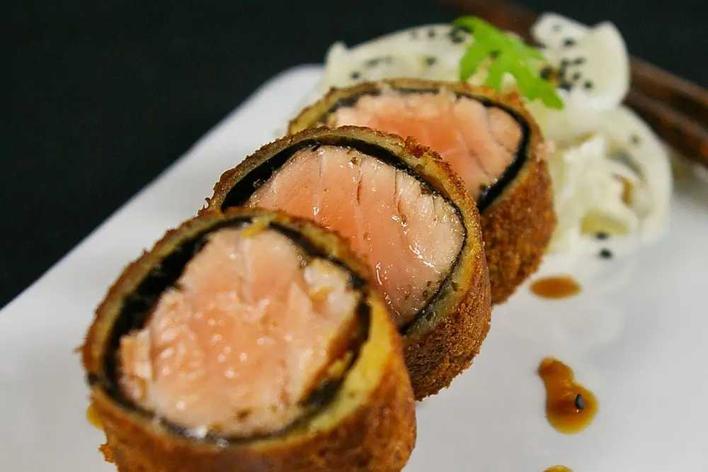 Lachs im Pankomantel mit eingelegtem Fenchel Lachs in ein Noriblatt gewickelt und anschließend in Panko paniert. In Scheiben Geschnitten und auf einer länglichen weißen Platte mit eingelegtem Fenchel und etwas Soße angerichtet.