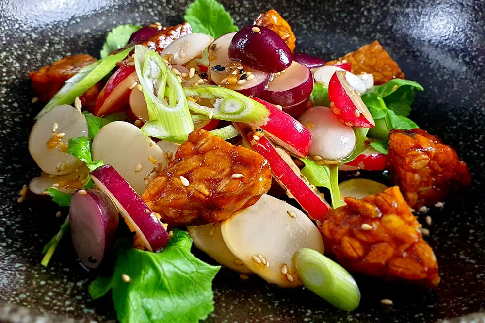 Radieschensalat mit Tempeh Farbenfroher frühlingshafter Salat mit bunten Radieschen und Krossem Tofu, garniert mit Frühlingszwiebeln. Angerichtet auf einem schwarzen tiefen Teller