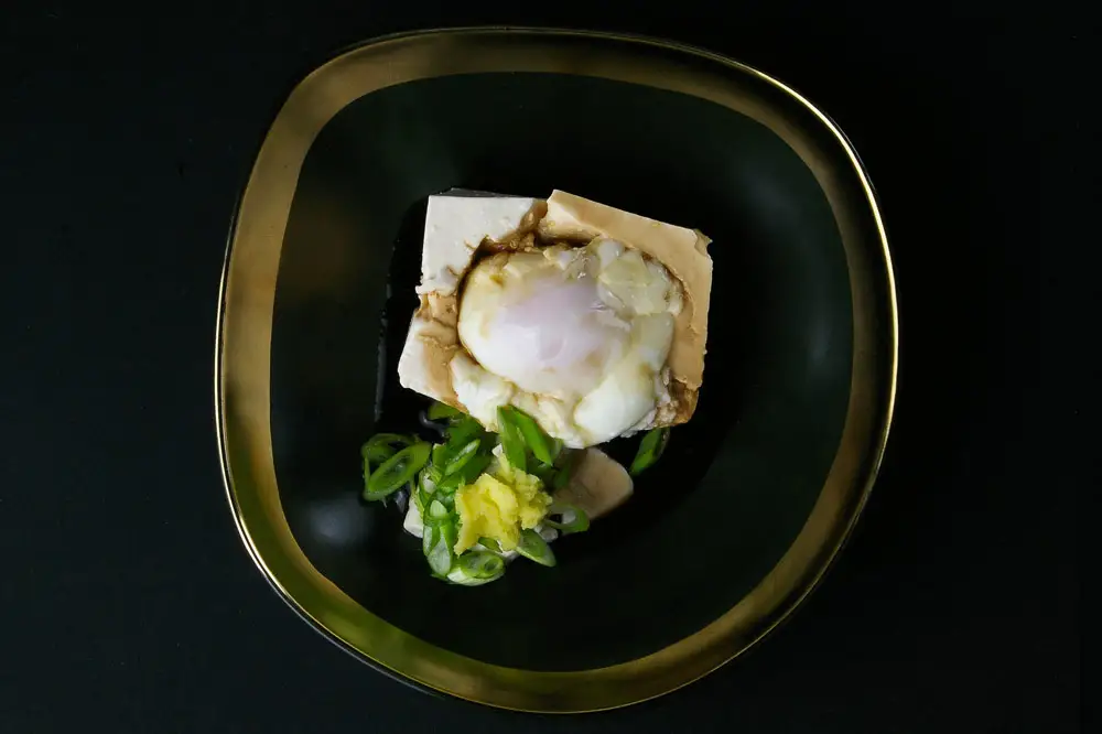 Seitentofu mit wachsweichem Ei Wunderbar feiner Seidentofu mit einem wachsweichen Ei, Frühlingszwiebeln Und einer würzigen Soße. In einem schönen schwarzen tiefen Teller mit Goldrand angerichtet.