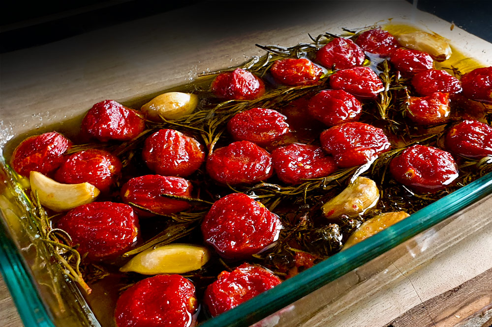 Confierte Tomaten, in einer Auflaufform liegen rote Cocktailtomaten mit Kräutern und Knoblauch, langsam gegart in Olivenöl.