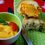 Tomatenbutter, hergestellt aus den Tomaten von meinem Rezept „confierte Tomaten“ und Butter. Angerichtet auf einem roten Teller mit grüner Serviette, sieht die wunderschöne orangefarbene Tomatenbutter in ihrem grünen Töpfchen, wunderschön aus.